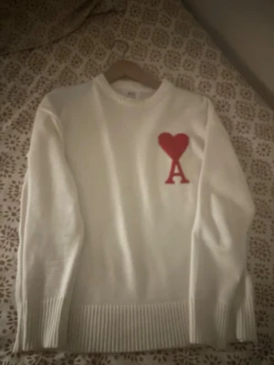 Vit stickad tröja från Ami Paris - Säljer en vit stickad tröja från Ami Paris med röd hjärtlogga och bokstaven A på bröstet. Tröjan har rund hals, ribbade muddar och är långärmad. Perfekt för dig som gillar stilrena plagg med en twist.