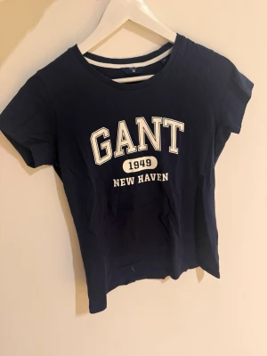 Mörkblå GANT t-shirt med tryck - Snygg mörkblå t-shirt från GANT med klassiskt tryck framtill där det står 'GANT 1949 NEW HAVEN'. T-shirten har rund halsringning och korta ärmar. Perfekt för en avslappnad look.