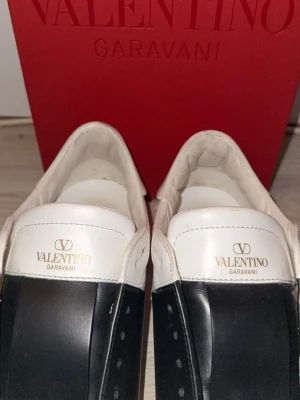 Valentino Garavani  - Säljer ett par snygga Valentino Garavani sneakers i vitt skinn med bred svart detalj över mitten. Klassisk låg modell med rund tå och platt sula. Kommer med originalkartong, dustbags och extra skosnören. Perfekta för dig som gillar exklusiva och stilrena sneakers. Allt medföljer såson skosnören, dustbags, kvitto från merchsweden och skulle det finnas frågor så är det bara att fråga!