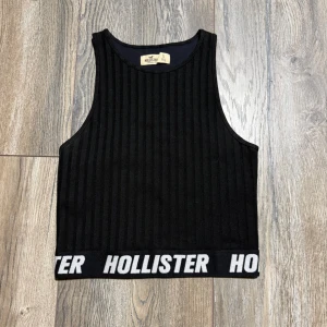Svart ribbad linnetopp Hollister - Säljer en svart ribbad linnetopp från Hollister i storlek S. Toppen är croppad och har breda axelband samt ett elastiskt band nertill med vit HOLLISTER-logga runt om. Perfekt till jeans eller shorts för en sportig look.