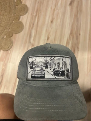 Grå meshkeps från Emilio Pedoniza - Säljer en grå keps från Emilio Pedoniza med mesh på baksidan och justerbar snapback. Framsidan har en broderad svartvit bild av en gata med bil och café. Kepsen har mjuk mockaliknande känsla framtill och är riktigt clean. Om du vill pruta är det bara skriva till mig 