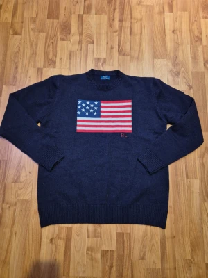 Polo Ralph Lauren - Dark blue knitted sweater from Polo Ralph Lauren with American flag embroidered on the chest and RL detail in red. Classic round neckline and ribbed cuffs at the sleeves and hem. Size is L
