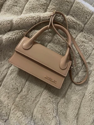Beige Jacquemus axelväska i skinn - Säljer en beige axelväska från Jacquemus i slätt skinn. Väskan har ett stilrent och minimalistiskt uttryck med guldfärgad logga framtill, topphandtag och justerbar axelrem. Perfekt storlek för det viktigaste och en riktig statement piece.