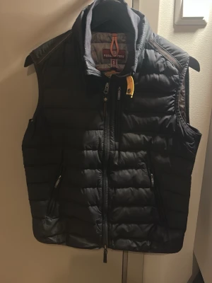 Svart dunväst från Parajumpers S - Säljer en svart dunväst från Parajumpers i storlek S. Västen är quiltad och har dragkedja framtill samt två fickor med dragkedja. Detalj med gul rem vid kragen och broderad logga på ryggen. Superlätt och perfekt för lager-på-lager.