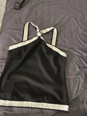 Svart och vit linnetopp med knyt - Säljer en svart linnetopp med vita kontrastband och snygg knytning i halsen. Toppen har raka linjer och breda axelband som möts i en knut framtill. Perfekt till jeans eller kjol för en clean look.