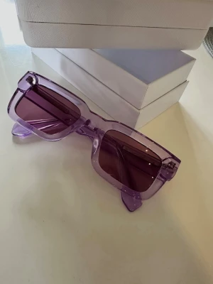 Lila solglasögon från CHIMI - Säljer ett par trendiga solglasögon från CHIMI i lila transparent plast med rektangulära bågar. Glasen är tonade i matchande lila nyans och bågarna har en cool, chunky design. Perfekt accessoar för att lyfta din stil i sommar!