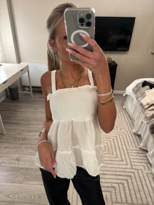 Vit volangig linnetopp - Supersöt vit linnetopp med breda axelband och smockad överdel. Toppen har volanger i flera lager och en luftig, flowy passform som känns perfekt för varma dagar. Tillverkad i ett tunt och lätt bomullstyg.