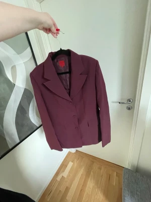 Vinröd kavaj  - En vinröd kavaj från Jacqueline De Young med klassisk krage och knappar framtill. Storlek 42