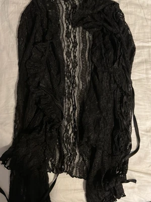 Svart spetskofta med snörning - Säljer en svart kofta i spets med snygga volangdetaljer och snörning framtill. Koftan har ett transparent, mönstrat tyg som ger en cool och unik look. Perfekt för att lyfta vilken outfit som helst.