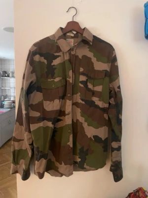 Camo skjorta från Vaillent Clothing - Säljer en camouflagemönstrad skjorta från Vaillent Clothing i storlek Large. Skjortan har klassisk krage, två bröstfickor med lock och knappar framtill. Tillverkad i bomull och har lång ärm. Perfekt för dig som gillar streetwear vibes.