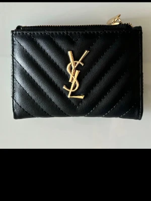 Svart quiltad plånbok från YSL - Snygg svart plånbok i quiltat skinn med guldiga detaljer och logotyp framtill. Plånboken har dragkedja upptill, flera kortfack och ett myntfack med tryckknapp. Perfekt för dig som gillar stilrena accessoarer med lyxig känsla. Helt oanvänd! Ny utan prislapp