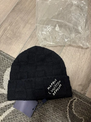 Svart Louis Vuitton mössa i kashmir - Säljer en svart mössa från Louis Vuitton med diskret rutmönster och broderad text framtill. Mössan är tillverkad i en lyxig mix av kashmir, ull och silke för extra mjuk känsla. Perfekt accessoar för dig som vill ha något stilrent och exklusivt.