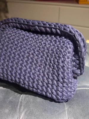 Mörkblå virkat clutch - Säljer en unik handväska i mörklila färg, virkad i ett tjockt garn som ger en cool och chunky look. Väskan har en rektangulär form och är öppen upptill, perfekt för att bära dina essentials. Perfekt accessoar för dig som gillar handgjorda detaljer.