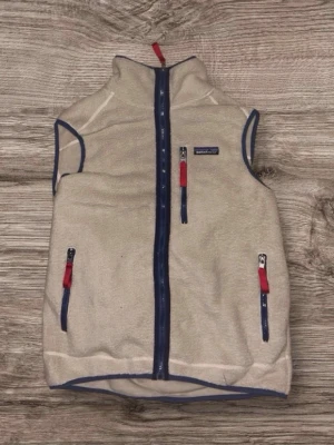 Beige fleeceväst från Patagonia S - Säljer en beige fleeceväst från Patagonia i storlek S. Västen har blå detaljer, hel dragkedja framtill och tre fickor med dragkedja och röda zip-pullers. Tillverkad i 100% ekologisk bomull och har en mjuk, mysig känsla. Perfekt för lager-på-lager under kyliga dagar.