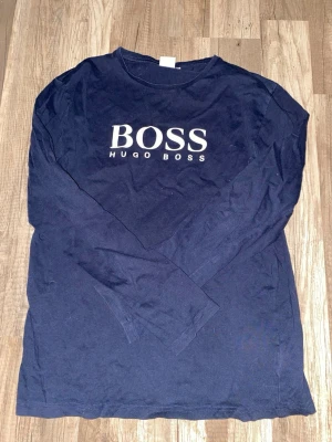 Mörkblå långärmad t-shirt Hugo Boss - Snygg mörkblå långärmad t-shirt från Hugo Boss med stor vit logga på bröstet. Klassisk rund halsringning och enkel design som funkar till det mesta. Tillverkad i mjuk bomull för skön känsla hela dagen.