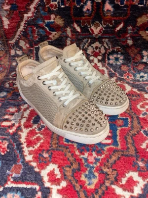 Christian Louboutin - Säljer ett par äkta beige sneakers från Christian Louboutin med coola silverfärgade nitar på tån. Skorna har snörning, meshpaneler på sidorna och detaljer i mocka. Snygg vit sula och rund tå. Perfekta för dig som vill sticka ut med din stil.