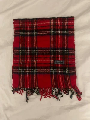Rutig röd halsduk från Burberrys - Säljer en klassisk rutig halsduk från Burberrys i rött, svart, vitt och gult. Halsduken är tillverkad i 100% lammull och har fransar i ändarna. Perfekt accessoar för att lyfta din outfit med en tidlös brittisk touch.