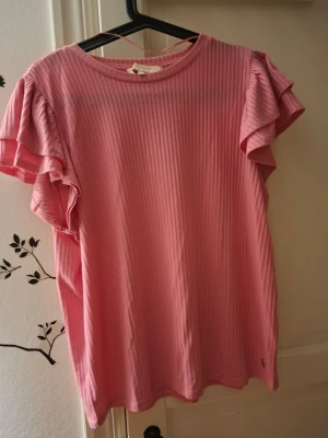 Rosa ribbad topp med volangärm - Säljer en söt rosa topp med ribbad struktur och volangdetaljer på ärmarna. Toppen har rund halsringning och korta ärmar med dubbla lager volang som ger en extra touch. Perfekt för dig som gillar en feminin och chill stil.