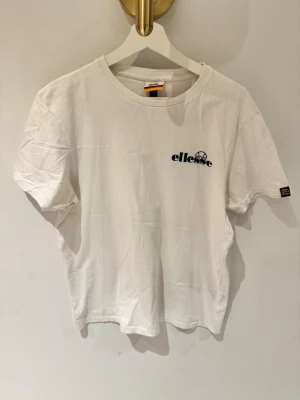 Vit Ellesse t-shirt med tryck - Säljer en vit t-shirt från Ellesse i mjuk bomull. Framsidan har en liten Ellesse-logga på bröstet och ärmen, medan baksidan har ett stort grafiskt tryck i svart, rött och grått. Klassisk passform och rund hals.