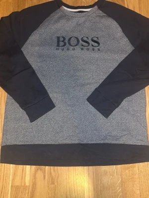 Grå och svart sweatshirt från Hugo Boss - Säljer en stilren sweatshirt från Hugo Boss i grått med svarta ärmar och muddar. Tröjan har stor BOSS-logga framtill och är tillverkad i mjuk bomullsmix. Perfekt för dig som gillar en clean och sportig look.