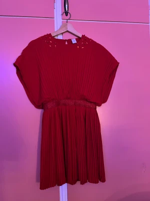 Röd plisserad klänning från Lindex - Superfin röd plisserad klänning från Lindex med glittrigt midjeband och dekorativa pärlor och stenar vid halsringningen. Klänningen har kort ärm och ett flowy fall som ger en snygg siluett. Perfekt för dig som vill sticka ut med färg och detaljer!