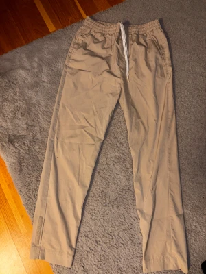 Beige mjukisbyxor från Lacoste - Snygga beige mjukisbyxor från Lacoste med elastisk midja och vit snörning. Byxorna har raka ben, sidofickor och diskret Lacoste-logga vid benslutet. Perfekta för chill dagar och har en clean look som funkar till sneakers.