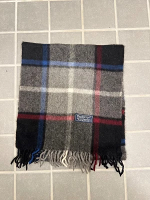 Rutig ullhalsduk från Burberrys - Klassisk rutig halsduk från Burberrys i 100% ull. Halsduken har fransar i ändarna och den ikoniska Burberrys-etiketten. Perfekt accessoar för att lyfta din outfit. Anledningen att de stör ”burberrys” är för att de är en äldre modell.