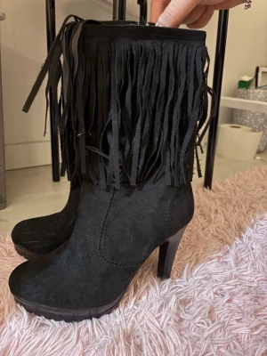Svarta boots med fransar och klack - Säljer ett par svarta boots med höga klackar och platt sula. Skorna har snygga fransdetaljer upptill. Dragkedja på sidan gör dem enkla att ta på. Perfekta för dig som vill sticka ut med cool western-vibe.