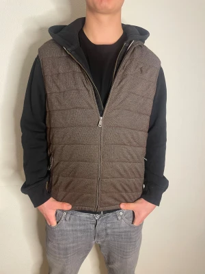 Brun väst från Polo Ralph Lauren - Snygg brun väst från Polo Ralph Lauren med quiltad design och broderad logga på bröstet. Västen har dragkedja framtill och två sidofickor med dragkedja. Materialet är polyester med en lätt textur som ger en cool look. Perfekt att slänga över en hoodie. Modellen är 188cm!