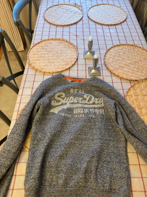 Grå långärmad Superdry tröja - Säljer en gråmelerad crewneck från Superdry med stort tryck på bröstet i vitt och japanska tecken. Tröjan har ribbade muddar vid ärmslut och nederkant samt en klassisk rund hals. Perfekt för dig som gillar streetwear och snygga loggor.