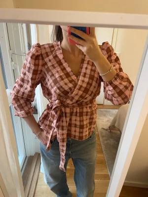 Rutig omlottblus med puffärm - Säljer en rutig omlottblus i rosa och brunt med puffiga axlar och långa ärmar. Blusen har knytband i midjan och ett mjukt, krinklat bomullstyg som ger en chill vibe. Perfekt för dig som gillar statement-ärmar och snygga detaljer.