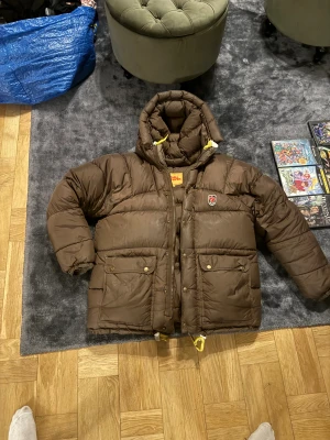 Brun Fjällräven dunjacka XXS - Säljer en brun dunjacka från Fjällräven i storlek XXS. Jackan har en rejäl huva med gula snören, två stora fickor med knappar och klassisk Fjällräven-logga på bröstet. Puffig och varm design, perfekt för kalla dagar.