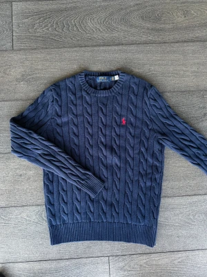 Marinblå stickad tröja Polo Ralph Lauren - Snygg marinblå stickad tröja från Polo Ralph Lauren med klassiskt kabelstickat mönster och röd broderad logga på bröstet. Rund hals och ribbade muddar vid ärmslut och nederkant. Perfekt för dig som gillar stilrena och tidlösa plagg.