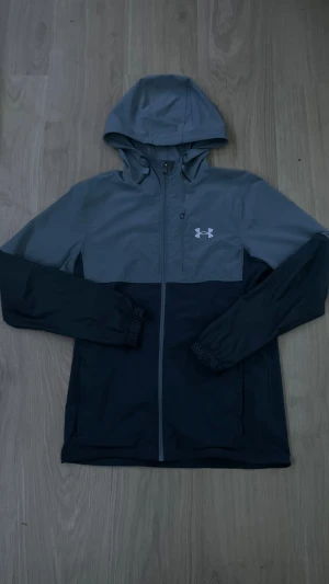 Grå och svart vindjacka Under Armour - Snygg tvåfärgad vindjacka från Under Armour i grått och svart. Jackan har huva, hel dragkedja framtill, lång ärm och en bröstficka med dragkedja. Perfekt för dig som gillar sportig stil och vill ha något lätt och funktionellt.