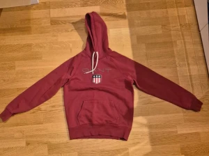 Vinröd hoodie från GANT med logga - Snygg vinröd hoodie från GANT med broderad logga och emblem på bröstet. Tröjan har huva med vita snören, känguruficka och ribbade muddar. Perfekt för dig som gillar klassisk stil med en sportig touch.