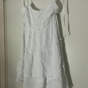 Vit somrig kortklänning med volanger - Luftig vit kortklänning med tunna axelband och volangdetaljer. Klänningen har en avslappnad passform och är tillverkad i ett lätt, krinklat tyg som ger en bohemisk känsla. Perfekt för varma dagar och enkel att styla med sandaler eller sneakers.