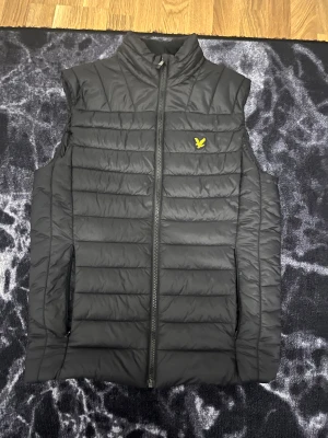 Svart dunväst från Lyle & Scott S - Svart dunväst från Lyle & Scott med gul broderad logga på bröstet. Västen har quiltad design, hög krage och dragkedja framtill. Tillverkad i polyester och har två sidofickor. Perfekt för lager-på-lager och snygg till streetwear.