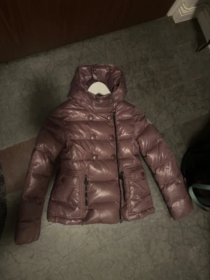 Lila pufferjacka med huva och knappar - Säljer en snygg lila pufferjacka med hög krage och huva. Jackan har dubbla rader med knappar framtill, dragkedja i sidan och två fickor med både knapp och dragkedja. Perfekt för kalla dagar och riktigt skön att bära.