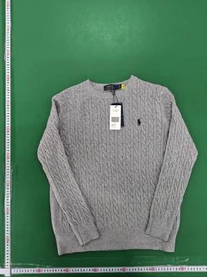 Grå kabelstickad tröja Polo Ralph Lauren - Snygg grå kabelstickad tröja från Polo Ralph Lauren i storlek L. Tröjan har rund hals, långärmad design och klassisk broderad logga på bröstet. Tillverkad i mjuk bomull som känns skön mot huden och passar perfekt till lager-på-lager-stil.