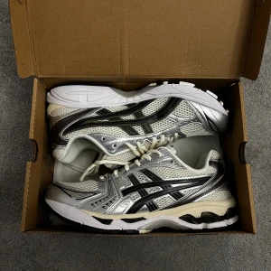 Asics Gel-Kayano 14 cream sneakers - Säljer ett par Asics Gel-Kayano 14 sneakers i silver och vitt med svarta detaljer. Skorna har mesh-överdel, syntetpaneler och snörning. Ikonisk retrodesign med platt sula och rund tå, perfekt för dig som gillar sportig stil och coola färgkombinationer.