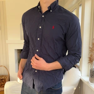 Mörkblå skjorta från Ralph Lauren - Snygg mörkblå skjorta från Ralph Lauren med klassisk passform och ikonisk röd logobrodering på bröstet. Skjortan har knappar hela vägen, button-down krage och långa ärmar. Tillverkad i mjuk bomull, perfekt för en clean och stilren look.