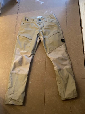 Beige cargopants från Stone Island - Snygga cargopants från Stone Island i beige med ljusare paneler på knän och lår. Byxorna har raka ben, flera fickor och klassisk Stone Island patch på sidan. Perfekt för dig som gillar streetwear och funktionella detaljer. Priset är ej hugget i sten😁