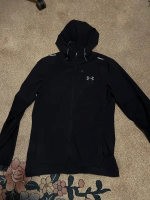 Svart vindjacka från Under Armour - Snygg svart vindjacka från Under Armour med huva och reflekterande detaljer. Jackan har dragkedja framtill, bröstficka med dragkedja och justerbar snörning vid huvan. Perfekt för dig som gillar sportig stil och vill ha något funktionellt. Litet hål finns som syns på bilden.