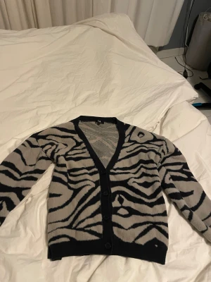 Zebramönstrad kofta från C&A - Snygg kofta från C&A i storlek M med zebramönster i svart och grått. Koftan har v-ringning, knappar framtill och långa ärmar. Mjuk stickad känsla som gör den skön att ha på sig. Perfekt för dig som gillar djurmönster och vill sticka ut lite extra.