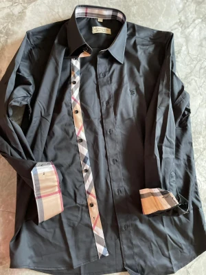 Svart Burberry skjorta - Svart skjorta från Burberry med klassiskt rutigt mönster på knappslå och insidan av manschetterna. Skjortan har lång ärm, knappar framtill och en snygg krage. Materialet är mjuk bomull och loggan är broderad på bröstet.