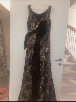 Svart långklänning med spets och rosett - Elegant svart långklänning med glittriga detaljer och spetsmönster. Klänningen har ärmlös design, stor svart rosett i midjan och dekorativa pärlor. Perfekt för dig som vill sticka ut med en glamorös look.