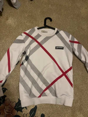 Vit rutig Burberry tröja S - Säljer en snygg vit tröja från Burberry i storlek S med klassiskt rutigt mönster i grått och rött. Tröjan har rund hals, lång ärm och Burberry-logga på bröstet. Perfekt för dig som gillar stilrena designer och vill sticka ut med ett ikoniskt märke.
