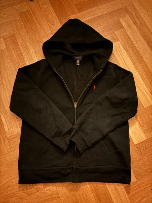 Svart hoodie från Polo Ralph Lauren XL - Svart hoodie från Polo Ralph Lauren i storlek XL. Tröjan har huva, dragkedja framtill och ett litet rött Polo-logobroderi på bröstet. Tillverkad i mjuk bomull med långärmad passform och två fickor framtill. Perfekt för chill dagar och streetwear.
