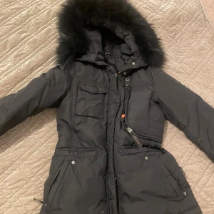 Svart dunjacka med päls från Hollies - Säljer en svart dunjacka från Hollies med fluffig päls på huvan, flera fickor med dragkedja och snygga detaljer. Jackan har lång ärm, markerad midja och quiltad design. 