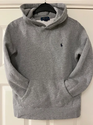 Grå hoodie från Polo Ralph Lauren - Snygg grå hoodie från Polo Ralph Lauren med klassisk huva och känguruficka framtill. Tröjan har lång ärm och ett diskret mörkblått Polo-logo broderat på bröstet. Perfekt för chill dagar och enkel att matcha med jeans eller joggers.